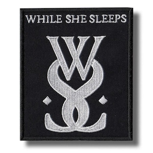 Puede incluir: Parche cuadrado negro con el texto "WHILE SHE SLEEPS" sobre un diseño estilizado plateado de "W" y "S". Dos pequeños diamantes plateados flanquean el diseño. El parche tiene un borde negro.