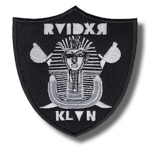 Könnte beinhalten: Schwarzer schildförmiger Aufnäher mit weißem und grauem Stickdesign. Das Design zeigt einen Pharaonenkopf, gekreuzte Äxte und die Texte "RVIDXR" und "KLVN".