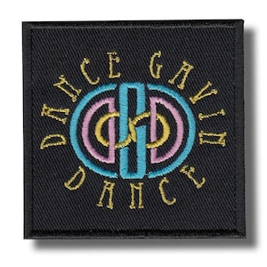 May include: Black square patch with embroidered text and design. The design features a central teal and gold shape, surrounded by a pink and gold double circle. The words "ICE GAVIN DANCE" are embroidered in gold around the circle.