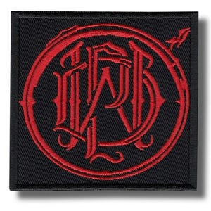 May include: Square black patch with a red, ornate monogram design. The monogram is encircled by a decorative border. The patch has a textured appearance and is likely made of fabric.