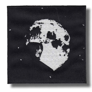Puede incluir: Parche cuadrado negro con un diseño bordado en blanco de una luna llena. La superficie de la luna está detallada con cráteres y sombras. Pequeños puntos blancos están dispersos alrededor de la luna, creando un efecto de cielo nocturno. El parche tiene un borde negro.