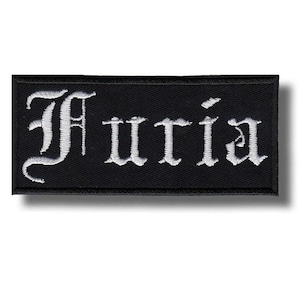 May include: A black rectangular patch featuring the word "Furia" in white gothic script. The patch has a black border and a shadow effect, giving a raised appearance.