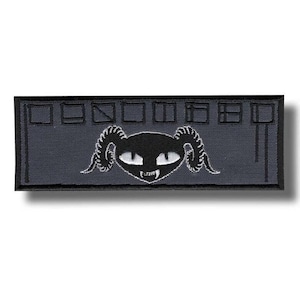 May include: A rectangular, dark gray patch featuring a black cat-like creature with horns, fangs, and white eyes. The creature is centered below a series of rectangular shapes. The patch is likely designed to be sewn or ironed onto fabric.