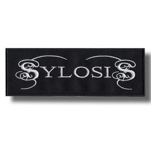 May include: A black rectangular patch with the word "SYLOSIS" embroidered in white, stylized with decorative flourishes. The patch is likely for clothing or accessories.