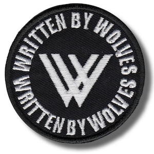May include: A round, black patch with white embroidered text and a white graphic. The text around the edge reads "WRITTEN BY WOLVES." Inside the circle is a stylized "W" graphic. The patch has a black border.