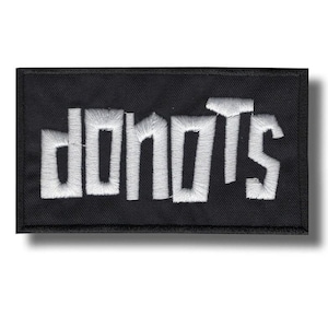 May include: A black rectangular patch with the word "DONUTS" embroidered in white, stylized block letters. The patch has a textured appearance and a shadow effect.