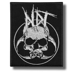 May include: Black embroidered patch featuring a white skull design within a circle. The skull has detailed features and is surrounded by a stylized, jagged crown and the letters "NAT". The patch is rectangular with a black border.