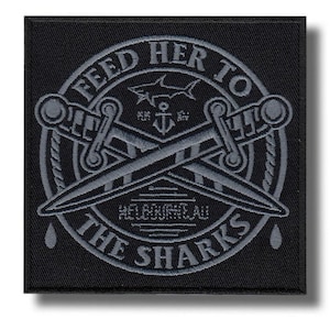 Peut inclure: Écusson carré noir avec texte et motif gris brodés. Le texte se lit "FEED HER TO THE SHARKS" autour d'un emblème central d'épées croisées, d'un requin et d'une ancre. Le mot "MELBOURNEAU" est également présent.