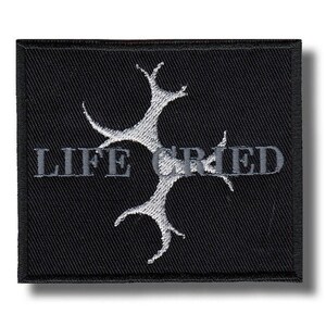 Pode incluir: Patch quadrado preto com as palavras "LIFE CRIED" bordadas em cinza, juntamente com um design de árvore estilizado e abstrato no centro. O patch tem uma borda preta e é provavelmente um patch para passar a ferro ou costurar.