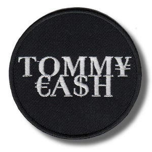 Op de afbeelding: Een ronde, zwarte geborduurde patch met de witte tekst "TOMMY CASH" en valutasymbolen. De patch heeft een zwarte rand en is waarschijnlijk ontworpen om op kleding of accessoires te worden genaaid of gestreken.