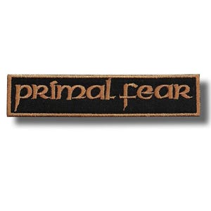 May include: A rectangular black patch embroidered with the words "primal fear" in gold thread. The patch has a gold border. It is designed to be sewn onto clothing or accessories, such as a jacket or bag.