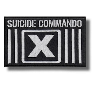 May include: Black rectangular patch with white embroidered text "SUICIDE COMMANDO". The patch features a white "X" inside a white square, flanked by vertical white stripes. The patch is likely for clothing or accessories.