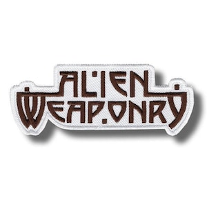 May include: Embroidered patch with the words "ALIEN WEAPON" in brown letters on a white background. The patch has a white border and a unique shape, with the text arranged in a stylized manner.