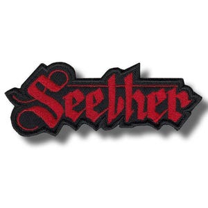 Puede incluir: Parche bordado con la palabra "Seether" en fuente gótica. El texto rojo está delineado en negro, sobre un fondo negro. El parche tiene un borde dentado.