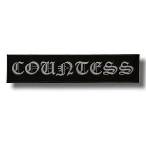 以下が含まれることがあります： 黒い長方形のパッチ。白いゴシック体で「COUNTESS」の文字が刺繍されています。パッチはシンプルでクリーンなデザインで、テキストは黒い背景の中央に配置されています。
