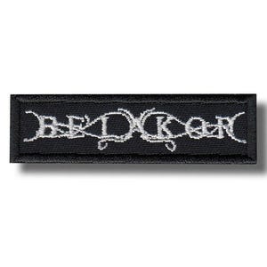 Op de afbeelding: Zwart rechthoekig embleem met wit geborduurde tekst. De tekst lijkt een bandnaam of logo te zijn. De patch heeft een zwarte rand en is waarschijnlijk gemaakt van stof. Het ontwerp is gothic of metal geïnspireerd.