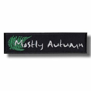 以下が含まれることがあります： 黒い長方形のパッチ。白い筆記体で「Mostly Autumn」と刺繍されています。パッチの左側には緑色のシダのデザインが刺繍されています。