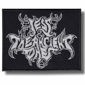 Pode incluir: Patch retangular preto com texto e símbolos bordados em branco. O texto é estilizado e gótico, com um design semelhante a um crânio. O patch é provavelmente para roupas ou acessórios, representando uma banda ou subcultura.