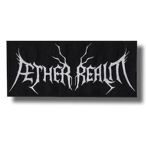 Pode incluir: Patch retangular preto com o texto branco "AETHER REALM" em uma fonte gótica estilizada. O texto tem serifs afiadas e pontiagudas e está centralizado no patch. O patch é provavelmente feito de tecido e foi projetado para ser costurado ou passado a ferro em roupas ou acessórios.