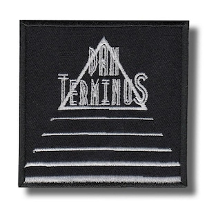 Puede incluir: Parche cuadrado negro con texto y diseño bordados en blanco. El texto dice "DAN TERMINUS" encima de una serie de líneas horizontales. El diseño incluye una forma de triángulo encima del texto. El parche es probablemente para ropa o accesorios.