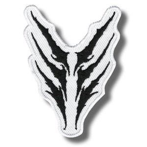 Peut inclure: Écusson brodé avec un motif noir et blanc. Le patch a un fond blanc avec un motif noir symétrique, ressemblant peut-être à un visage d'animal stylisé ou à un motif tribal. Les bords sont soigneusement cousus.