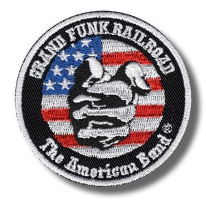 May include: Embroidered patch featuring the Grand Funk Railroad logo. The design includes a stylized fist over an American flag background, with the text "GRAND FUNK RAILROAD" and "The American Band" in a circular border.