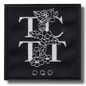 Op de afbeelding: Zwart vierkantige patch met witte geborduurde bloemen en de letters "TT" verticaal gestapeld. De patch heeft een zwarte rand en drie kleine witte cirkels aan de onderkant. Het ontwerp is gedetailleerd.