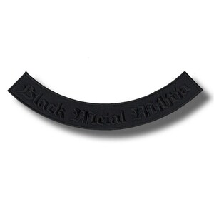 Pode incluir: Patch preto curvo com as palavras "Black Official Militia" em uma fonte gótica. O patch é de cor preta sólida e tem uma aparência em relevo texturizada. Provavelmente para uma jaqueta ou colete.