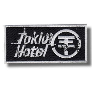 Puede incluir: Parche rectangular negro con texto bordado blanco y un símbolo. El texto dice "Tokio Hotel" en una fuente desgastada. Un círculo blanco con una "T" estilizada está a la derecha. El parche tiene un borde blanco.