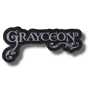 以下が含まれることがあります： 「GRAYCEON」の文字がスタイリッシュなライトグレーで刺繍された黒いパッチ。パッチには装飾的な縁取りがあり、衣類やアクセサリーに縫い付けるようにデザインされています。