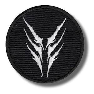 May include: A round, black embroidered patch featuring a white, stylized design. The design is symmetrical, with sharp, angular shapes radiating from the center. The patch has a black border and is likely for clothing or accessories.