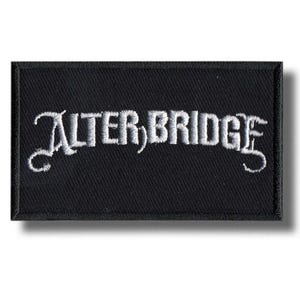 Puede incluir: Parche rectangular negro con el texto bordado "ALTER BRIDGE" en blanco. El texto está en una fuente estilizada. El parche tiene un borde negro y un efecto de sombra.