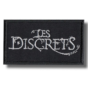 Puede incluir: Un parche rectangular negro con las palabras "LES SECRETS" bordadas en blanco. El diseño es limpio y minimalista, con el texto centrado y los bordes cuidadosamente acabados.