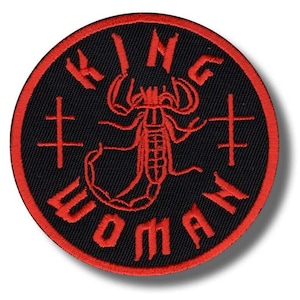 May include: A round, black embroidered patch with a red border. The patch features a red scorpion design in the center, surrounded by the words "KING WOMAN" in red, and four red cross symbols.