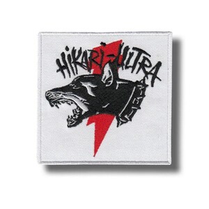 Op de afbeelding: Vierkantige witte patch met een zwarte hondenkopafbeelding, een rode bliksemschicht en de tekst "HIKARI ULTRA" in zwart. De hond heeft een open bek en een halsband met studs. De patch is voor kleding of accessoires.