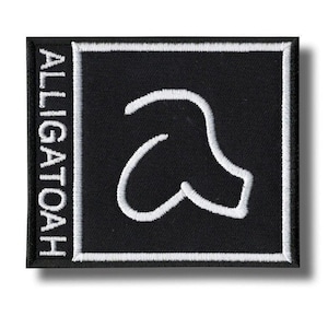 Op de afbeelding: Zwart vierkantige patch met witte geborduurde rand en een witte gestileerde letter "a". Het woord "ALLIGATOAH" is verticaal in wit geborduurd aan de linkerkant. Geschikt voor kleding of accessoires.