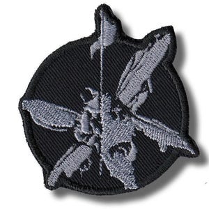 Pode incluir: Patch bordado com um design estilizado de inseto cinza sobre um fundo circular preto. O inseto tem asas e corpo detalhados, com um elemento em forma de bandeira na parte superior. O patch tem uma borda preta.