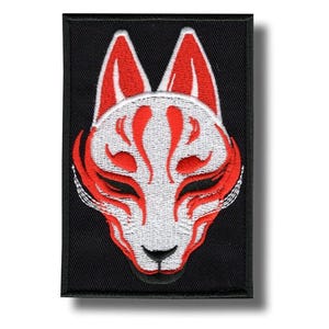 Pode incluir: Patch bordado com um design de máscara de raposa branca estilizada com detalhes vermelhos sobre um fundo preto. O design da máscara de raposa inclui detalhes vermelhos ao redor dos olhos, nariz e boca, e nas orelhas. O patch é retangular.