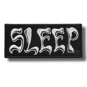 May include: Black rectangular patch featuring the word "SLEEP" in white, ornate embroidery. The patch has a black border and is designed to be sewn onto clothing or accessories. The design is simple but striking.