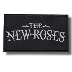 Könnte beinhalten: Schwarzer rechteckiger Aufnäher mit dem gestickten Text "THE NEW ROSES" in Weiß, in stilisierten Buchstaben. Der Aufnäher hat einen schwarzen Rand und ist wahrscheinlich zum Aufnähen auf Kleidung oder Accessoires gedacht.