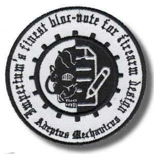 Op de afbeelding: Een cirkelvormige geborduurde patch met een zwart-wit ontwerp. De centrale afbeelding toont een schedel, een notitieblok en een potlood. De buitenste ring heeft tekst in een gotisch lettertype en een tandwielachtige rand.
