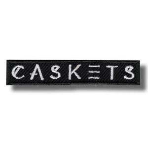 以下が含まれることがあります： 黒い長方形のパッチ。白い糸で「CASKETS」の文字が刺繍されています。文字ははっきりとしており、パッチはすっきりとしたシンプルなデザインです。