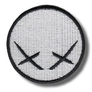 Può includere: Una toppa circolare con sfondo bianco e bordo nero. Due forme di 'X' nere sono centrate sulla toppa. La toppa ha un aspetto strutturato.
