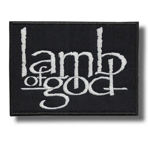 May include: Black rectangular patch with white embroidered text that reads "Lamb of God". The patch has a black border and a shadow effect, creating a 3D appearance. The text is in a stylized font.