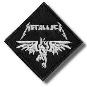Op de afbeelding: Zwart, ruitvormig embleem met witte borduursels. Het ontwerp toont de naam "METALLICA" boven een gestileerde gevleugelde figuur. Geschikt voor het aanbrengen op kleding of accessoires.