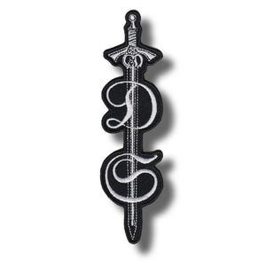 Pode incluir: Patch bordado a preto e branco com um desenho de espada e as letras "D" e "S" em uma fonte estilizada. O patch tem um fundo preto com costuras brancas, criando um design de alto contraste. A guarda e a lâmina da espada são detalhadas.