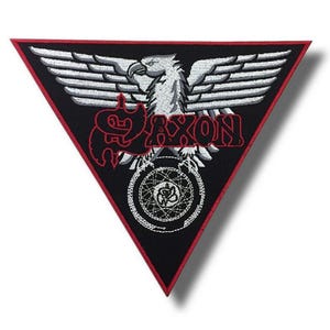 Może przedstawiać: Trójkątna czarna naszywka z czerwoną obwódką przedstawia białego orła z rozpostartymi skrzydłami. Słowo "SAXON" jest wyhaftowane na czerwono, a pod orłem znajduje się okrągły wzór. Ta naszywka jest prawdopodobnie przeznaczona na kurtkę lub torbę.