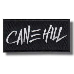 May include: Black rectangular patch featuring the white text "CANE HILL" in a stylized script. The patch has a textured appearance, and the text is embroidered. The patch is likely for clothing or accessories.