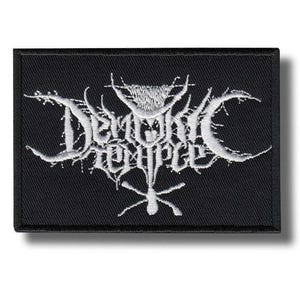 May include: Black rectangular patch with the white text "Demonic Temple" in a gothic font. The patch has a white embroidered design with a cross-like symbol at the bottom. The patch is likely for clothing or accessories.