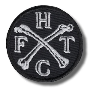 May include: Round black patch with white embroidered design. Two crossed bones are in the center, with the letters H, F, T, and C surrounding the bones. The patch has a stitched border.